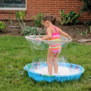 A girl in a kiddie pool using a hula hoop to create a large soap bubble